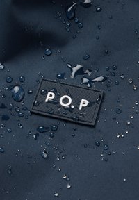 Vesitiivis tummansininen kangas, jossa on tekstuurikuvio; siinä on vesipisaroita ja suorakulmainen musta etiketti, jossa on valkoisilla kirjaimilla "P.O.P".