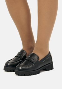 Mocassins en cuir noir avec des clous en argent portés avec des collants transparents couleur tan sur des jambes debout contre un fond blanc.