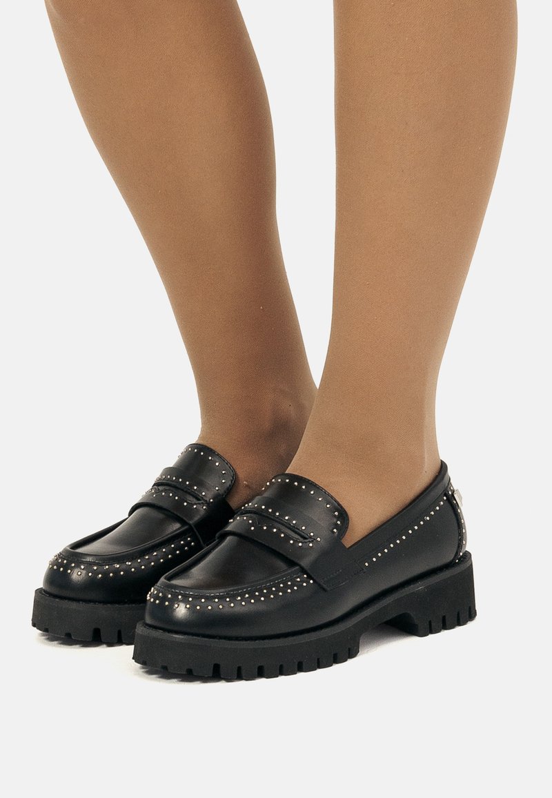 Mocassins en cuir noir avec des clous en argent portés avec des collants transparents couleur tan sur des jambes debout contre un fond blanc.