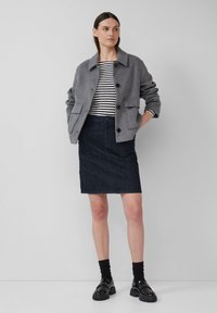 Veste en mélange de laine grise avec un col, deux poches avant, associée à une jupe en denim noire et des mocassins noirs sur des chaussettes noires.
