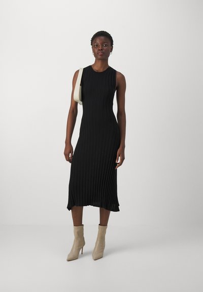 Robe midi noire côtelée sans manches, avec un ourlet arrondi. Le modèle porte des bottines à talons pointus beiges et un sac à épaule blanc cassé.