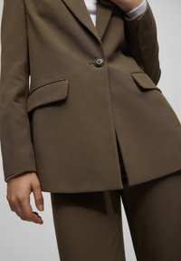 Brown tailored blazer with two front pockets, a single button closure, and a structured fit. Fabric has a smooth texture, slightly stretchable.