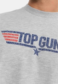 Šedé bavlněné tričko s modrou grafikou "TOP GUN" se zlomenou texturou a červeným pruhovaným designem křídel. Klasický kulatý výstřih.
