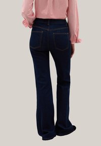Jeans de campana azul oscuro con una textura suave, que cuentan con dos bolsillos traseros y costuras en contraste. Llevados con una camisa de rayas de manga larga.