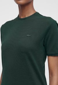 Gros plan d’une personne portant une chemise à manches courtes vert foncé avec des bords festonnés et un petit logo crocodile brodé sur la poitrine gauche.