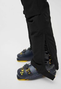 Bottes de ski grises avec des accents jaunes, boucles robustes, associées à un pantalon de ski noir avec un détail zippé sur le côté.