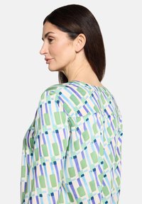 Bunte Bluse mit einem geometrischen Muster in Grün, Blau und Weiß. Hergestellt aus leichtem Stoff mit rundem Ausschnitt und rückseitigem Passe.