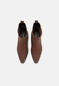 Botas de tobillo marrones con parte superior de tela suave, diseño de punta afilada y suelas de goma negras; presentan un pequeño lazo en la parte posterior para facilitar su uso.