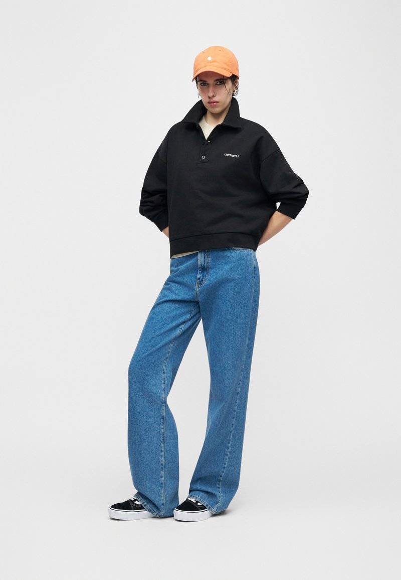 Jeune personne portant un sweat-shirt noir Carhartt, un jean bleu à jambes larges, des baskets noires et blanches, et une casquette de baseball orange, debout les mains sur les hanches.