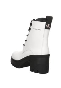 Botas tobilleras de cuero sintético blanco con suela gruesa negra, punta redonda y detalle de cordones. Presenta un logotipo en el tacón y un acento negro en la parte superior.