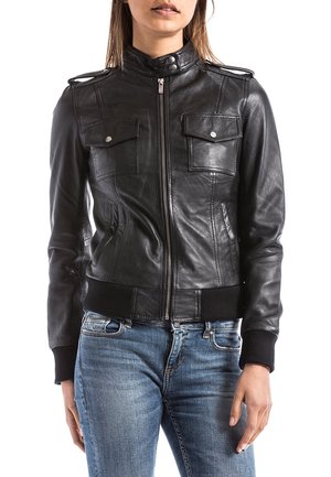 Femme portant une veste en cuir noire zippée avec deux poches avant à rabat et un jean bleu, debout, les mains détendues le long du corps.