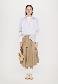 Cămașă oversized cu dungi albastre și albe, fustă bej cu fundă, șosete albe, adidași bej și o geantă țesută maro.