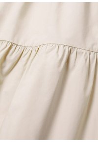 Gros plan d’un tissu beige avec des coutures froncées créant des plis doux, probablement partie d’un vêtement ou d’un rideau.