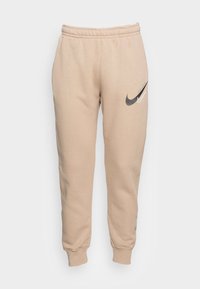 Béžové tepláky vyrobené z měkké látky, s elastickým pasem a zúženými manžetami, s tmavě šedým logem Nike na levé straně.