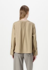 Chemisier beige à manches longues avec un col arrondi et un détail froncé à l'arrière ; tissu doux avec des poignets à bouton et une coupe décontractée.