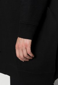 Hand with light skin tone resting against black long-sleeve garment with ribbed cuff, showing part of the arm and waist area.