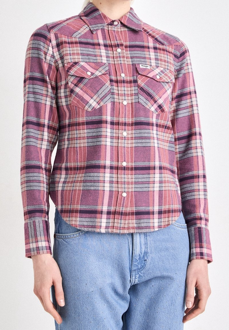 Personne portant une chemise à carreaux rose et violette boutonnée avec deux poches poitrine et un jean bleu clair sur fond blanc.