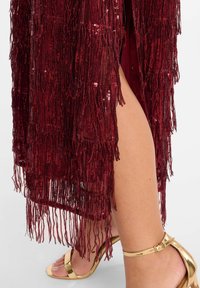 Bordeaux jurk met franjes en pailletten, voorzien van een zijsplit. Metallic gouden hak met enkelbandje zichtbaar onderaan.