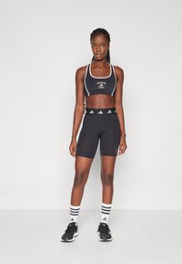 Černý sportovní crop top a šortky s vysokým pasem, zdobené bílým logem Adidas a designem se třemi pruhy na ponožkách, doplněné černými sportovními teniskami.