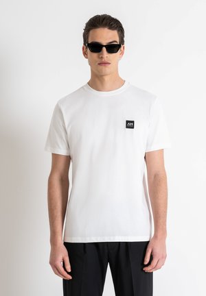 Camiseta de algodón blanca de manga corta, con escote redondo y un parche rectangular negro con el logotipo en el lado izquierdo del pecho. Se lleva con pantalones negros.