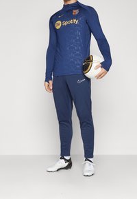Marineblauw sportshirt met lange mouwen en ritsen, met een gestructureerd patroon, logo en gouden accenten; gecombineerd met bijpassende broek en een voetbal.