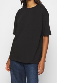 Person som bär en lös svart t-shirt med kort ärm och en liten logga på bröstet, samt blå denimjeans, stående mot en neutral bakgrund.