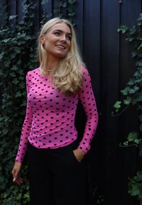 Rózsaszín hosszú ujjú felső fekete szívmintákkal, környakkivágással és testhez simuló szabással; fekete nadrággal párosítva, sötét fa háttér előtt.