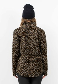 Luipaardprint fleece pullover, bruine en zwarte kleuren, kraag, lange mouwen, ronde zoom, zachte textuur. Draagt een zwarte beanie.