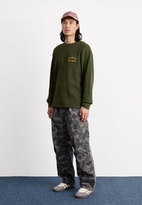 Suéter verde de manga larga con un acabado texturizado y texto amarillo; pantalones gris camuflados con bolsillos cargo; zapatos grises con franjas blancas; gorra rosa.