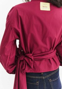 Bordowa bluzka kopertowa z długimi rękawami, ozdobiona dużą kokardą z tyłu oraz etykietą materiałową przyszytą przy kołnierzu. Wykonana z teksturowanej bawełny.