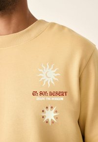 Beige sweatshirt med hvide og røde solgrafikker med teksten "EL SOL DESERT" og "CHASE THE HORIZON." Ribbet halsudskæring, blød tekstur.