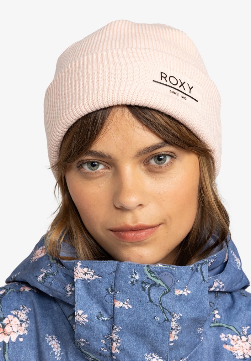 Světle růžová žebrovaná čepice s černým logem "ROXY", nošená s modrou bundou s květinovým vzorem, která má vysoký límec a zapínání na knoflíky.
