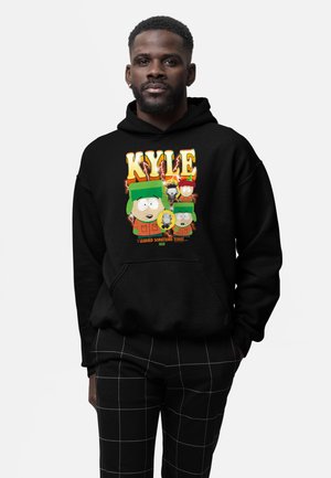 Uomo con felpa nera con cappuccio con la scritta "Kyle" e personaggi dei cartoni animati di South Park, abbinata a pantaloni neri con motivo a quadri bianchi.