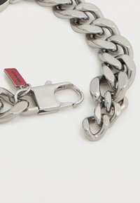 Βραχιόλι με μεταλλική αλυσίδα που διαθέτει μεγάλα, συνδεδεμένα κρίκους σε γυαλιστερό ασημί, με κούμπωμα και μία μικρή κόκκινη ετικέτα με την ένδειξη «HUGO».