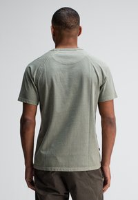 Man met kort haar, draagt een vervaagde groene T-shirt met korte mouwen en donkere broek, staande met zijn rug naar de camera tegen een effen achtergrond.