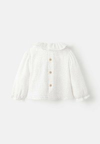 Witte blouse met een gerimpelde kraag, lange mouwen met elastische manchetten en vijf houten knopen op de rug, voorzien van kleine bloemenpatronen.