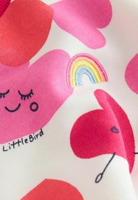 Ylhäällä kuvioitu paita, jossa on valkoinen tausta, suuret vaaleanpunaiset ja punaiset muodot, brodeerattu sateenkaari sekä hymyilevä kasvojen design; pehmeä tekstuuri.