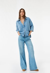 Lyseblå denimskjorte med knaplukning og oversized pasform, parret med matchende højtaljede, bredbenede jeans og brune sko.