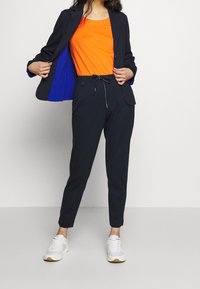Donna che indossa pantaloni e blazer blu scuro con una maglietta arancione brillante e sneakers bianche, mostrando l'imbottitura blu del blazer.