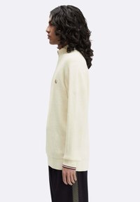 Maglione a maglia color crema con colletto alto, tessuto testurizzato e spacchi laterali. Presenta un piccolo logo ricamato sul petto e polsini a righe.