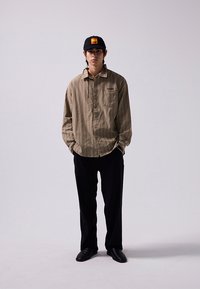 Brown striped button-up shirt, black pants, and black shoes. Black cap with orange logo. Model stands upright with hands in pockets.
