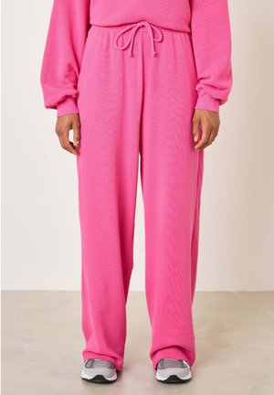 Pantalons de jogging roses avec taille à cordon, jambes larges et texture douce, associés à des chaussures de sport grises.