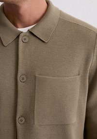 Khaki gebreide shirt met een kraag, twee knoopsluitingen en een opgestikte zak op de linkerborst. Glad textuur, lichtgewicht stof.