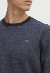 Donkerblauwe gebreide trui met een textuurpatroon, voorzien van een ronde halslijn en een klein cirkelvormig logo op de linkerkant van de borst.