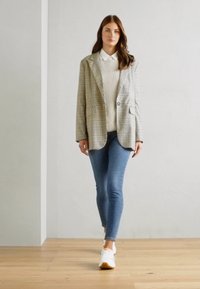 Rutig blazer i beige och grått över en krämfärgad tröja, ihop med blå skinny jeans och vita sneakers. Trägolvsbakgrund.