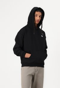 Schwarzer Kapuzenpullover aus Baumwollmischung, mit einer Fronttasche und einem kleinen Logoschild. Das Model trägt hellgraue Hosen.