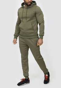 Survêtement vert olive avec capuche, poche kangourou et détail de logo. Design ajusté avec des poignets côtelés. Chaussures de sport noires et orange.