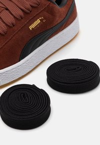 Puma Suede Sneaker aus rotbraunem Wildleder mit schwarzen Akzenten, ausgestattet mit einer strukturierten weißen Mittelsohle und zwei gerollten schwarzen Schnürsenkeln.