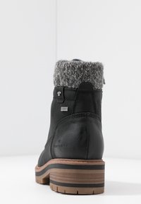 Bottine noire avec manchette tricotée grise, semelle texturée et talon empilé, vue de dos sur une surface blanche.