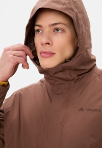 Veste imperméable marron avec un col haut et une capuche réglable, dotée d'une fermeture éclair et du logo "Vaude" en relief à l'avant.